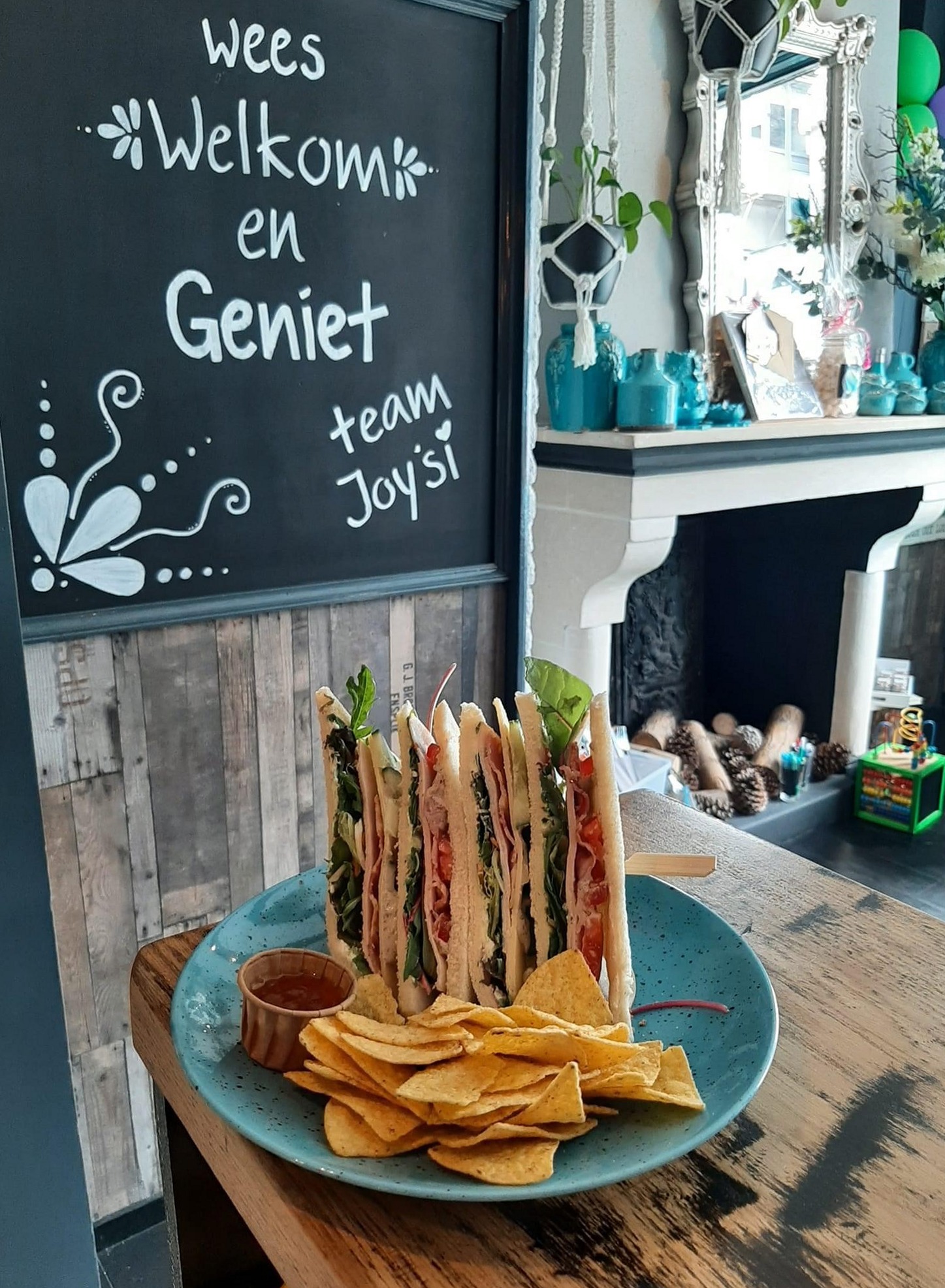 Een rijk belegd broodje met tortillachips naast het welkomstbord van Joy’Si